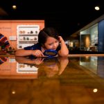 A young girl looks down through a magnifying glass at an antique map on display under glass.