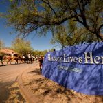 A wide-angle shot of historic reenactors on horseback riding down a road; in the foreground is a sign reading "History Lives Here."