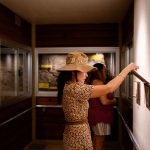 A young woman in a sundress and straw hat stands in an exhibit and examines an interactive flip panel.